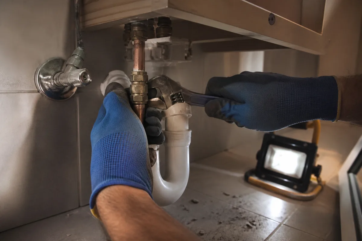Mains gantées raccordant un tuyau en cuivre sous un meuble de salle de bains, éclairage de chantier